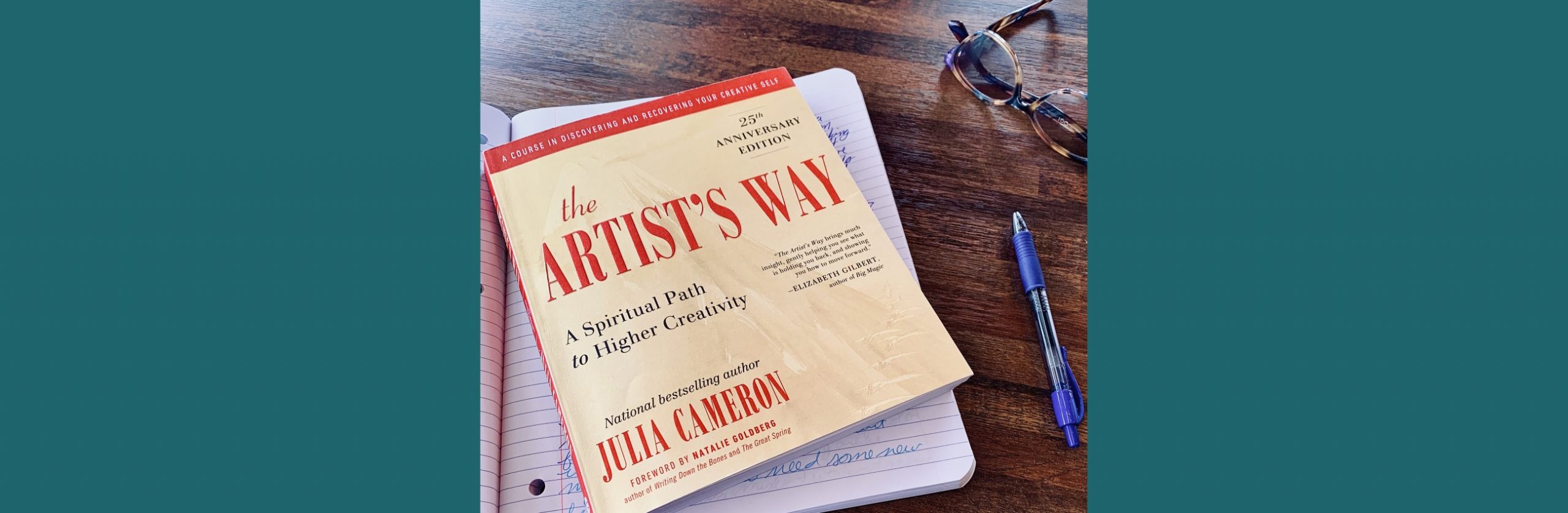 The Artist's Way book lays on an open notebook next to a blue pen and a pair of glasses