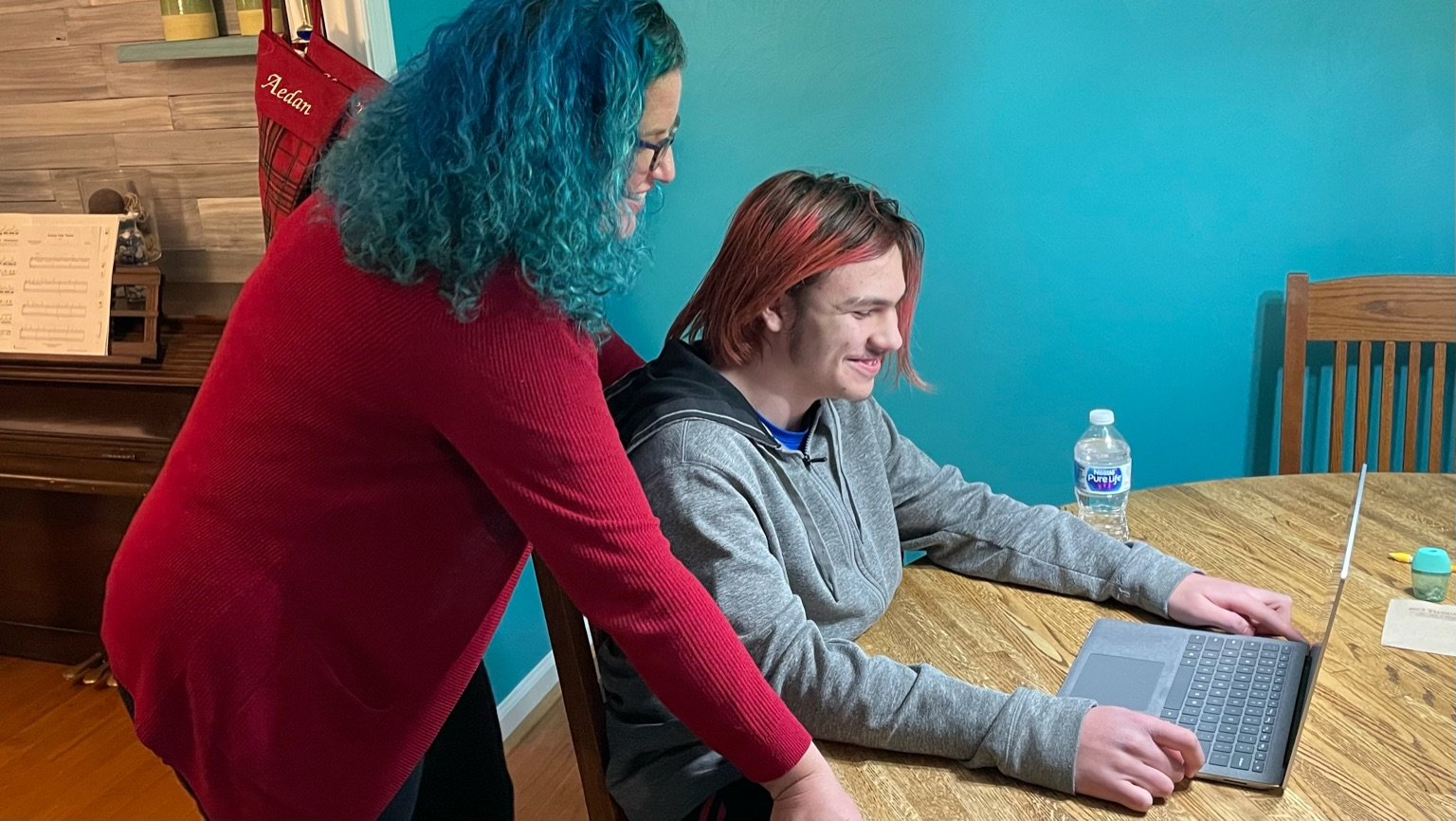 Pandemic parenting at its best: woman with blue hair and red sweater stands and leans over seated boy with long red hair who is smiling while looking at laptop on the table in front of him