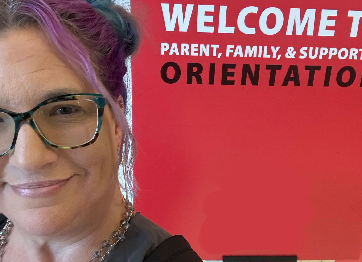 Kristen sporting space buns stands in front of red sign that says Welcome | Parent, Family, & Supporter Orientation