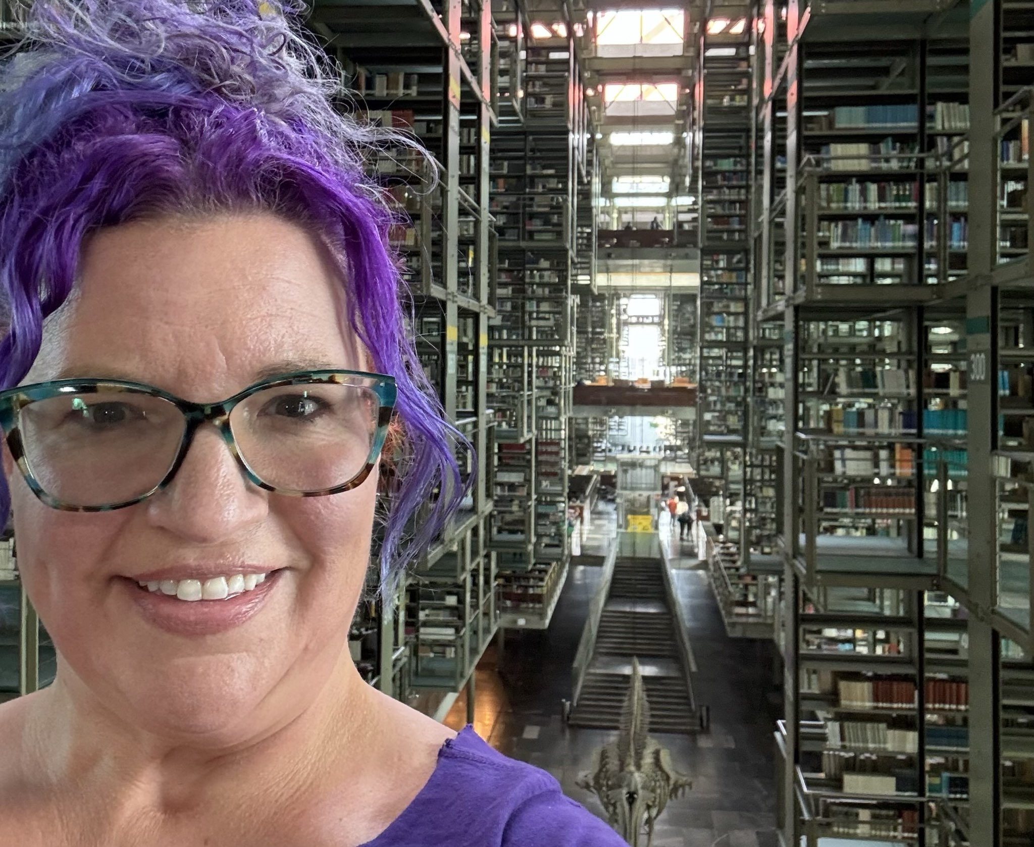 Kristen with purple hair in ponytail stands in front of shelves and shelves of books